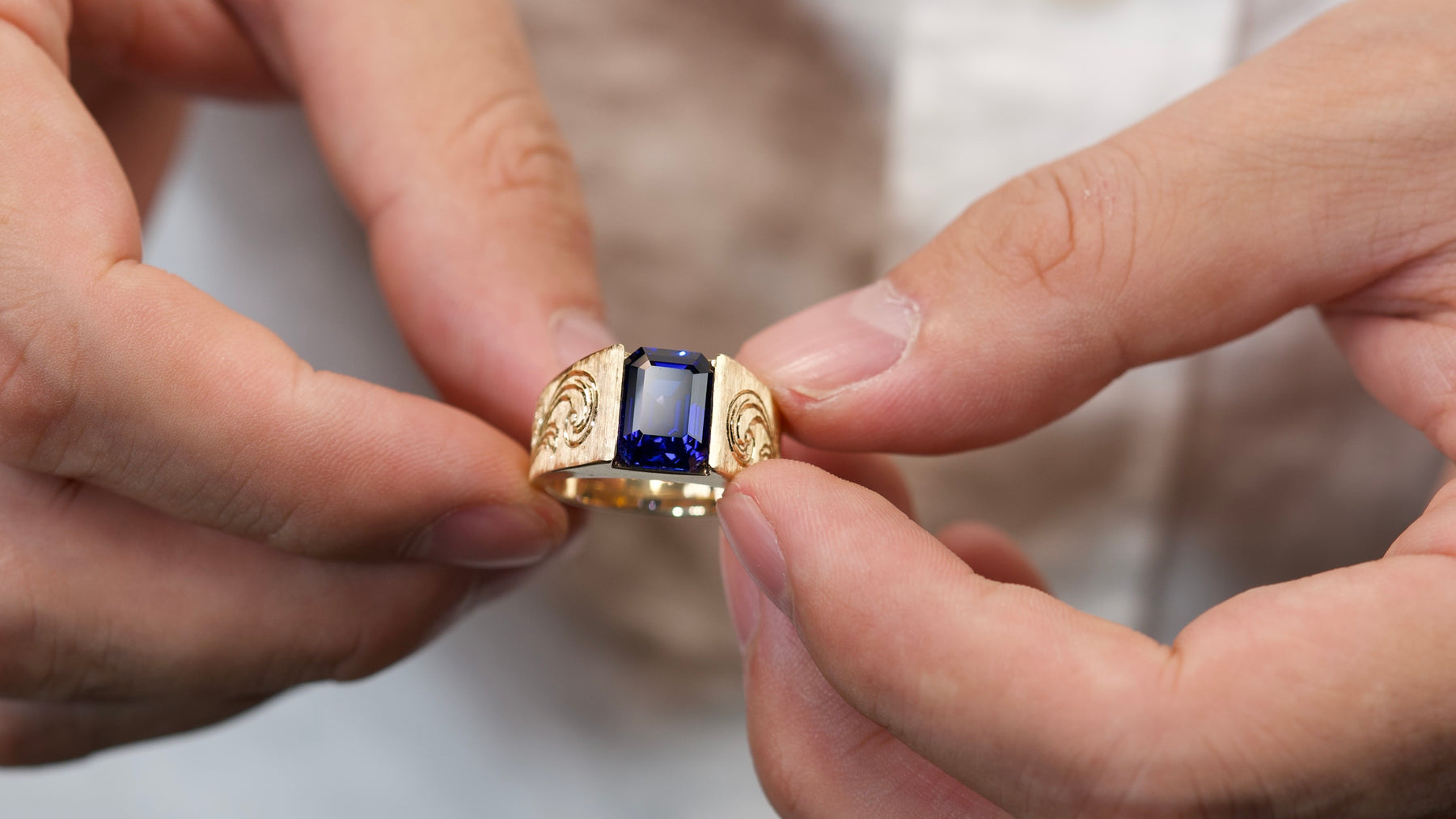 Hands holding a custom wide-band ring with a deep-blue rectangular center stone and engraved shoulders.
