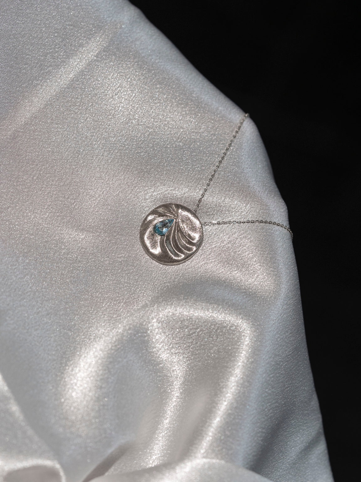 LIN 14k white gold pendant with silk-inspired folds and a pear-shaped aquamarine, shown on pale silk fabric.