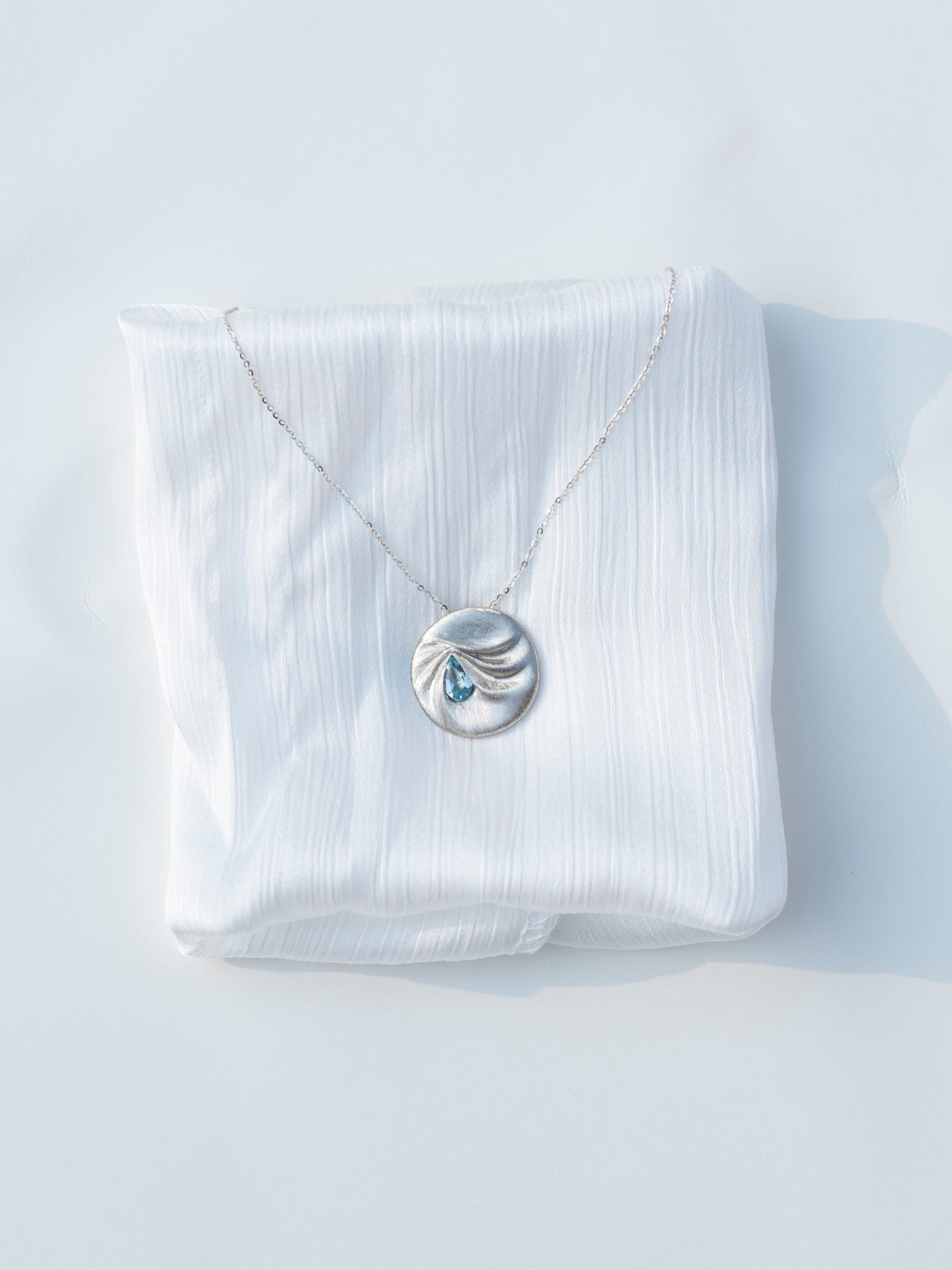 Front view of the LIN 14k white gold aquamarine pendant, displayed with a chain on folded white fabric.