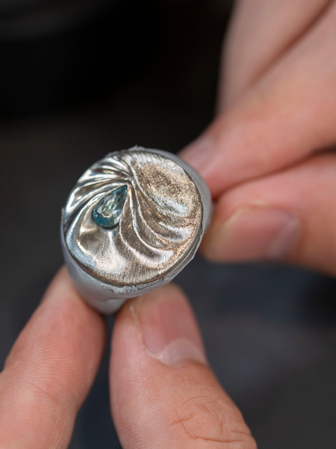 Close-up of LIN's 14k white gold aquamarine pendant on a work holder, showing the hand-worked surface texture and folds around the stone.