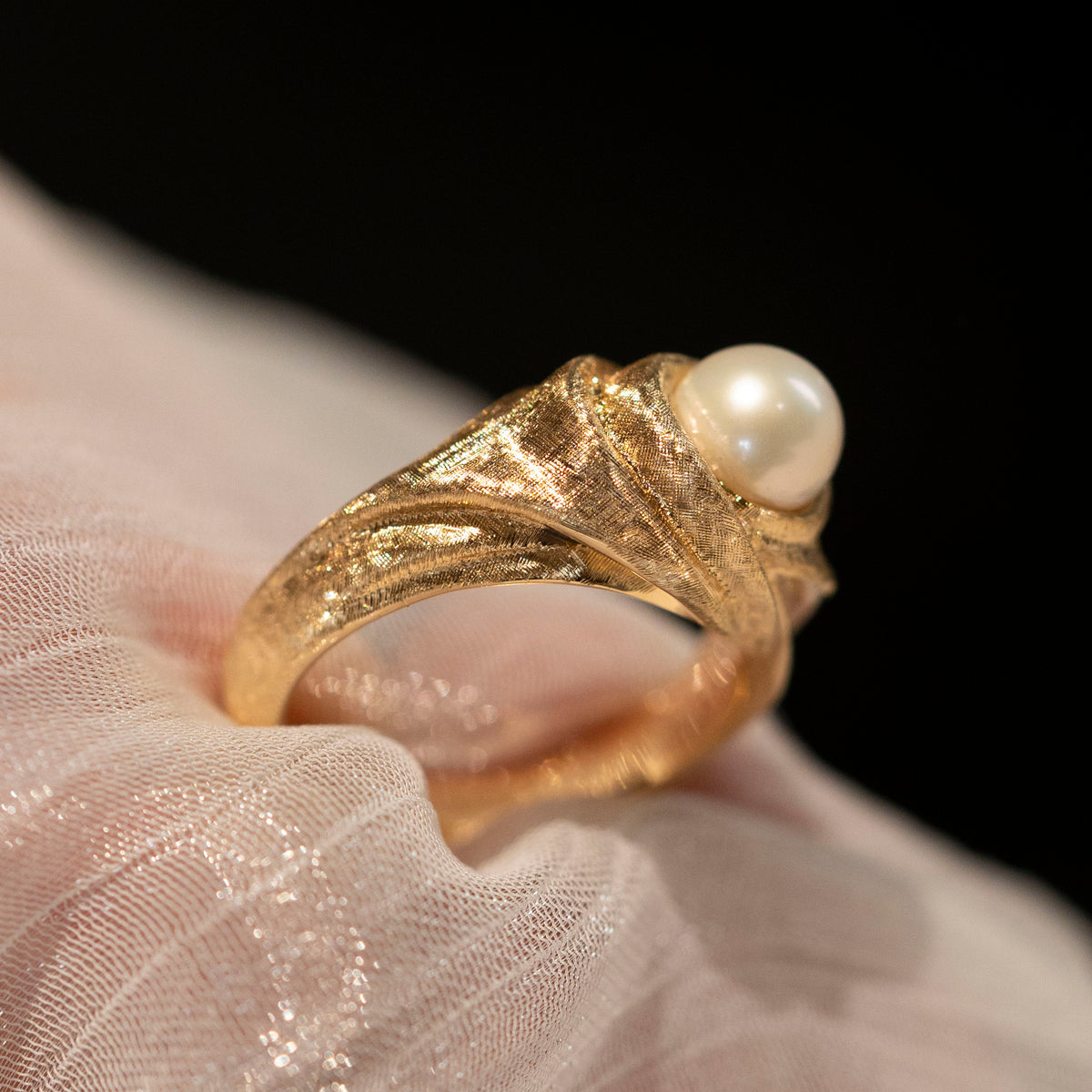 Side view of the 18K gold pearl ring, showing sculptural folds and the ring band on sheer fabric.